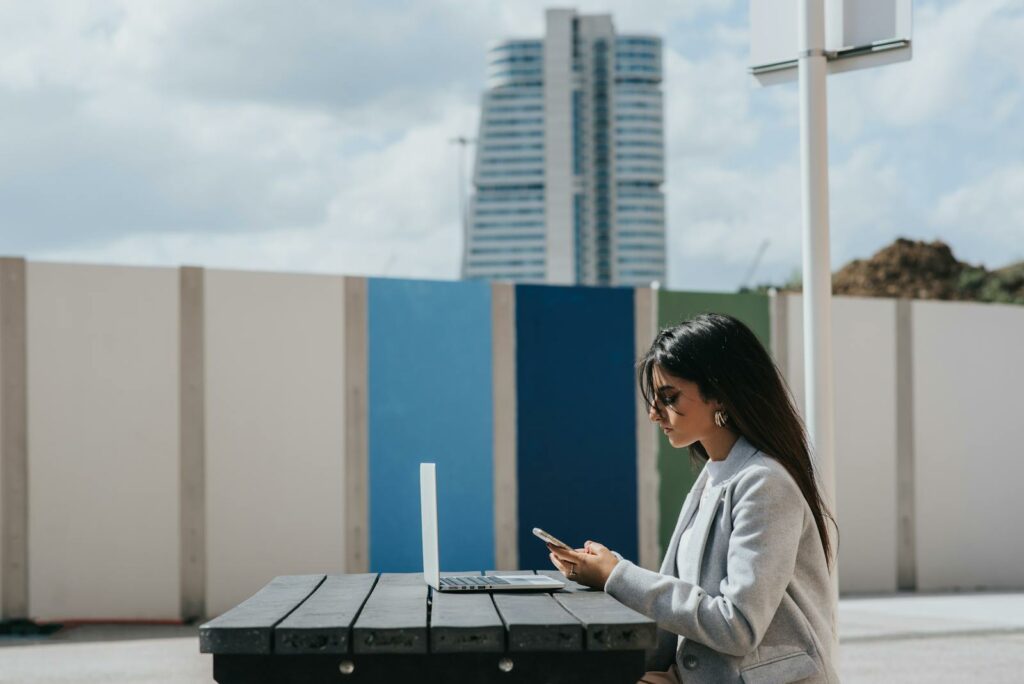 Ethnic female executive watching smartphone at table with laptop outdoors