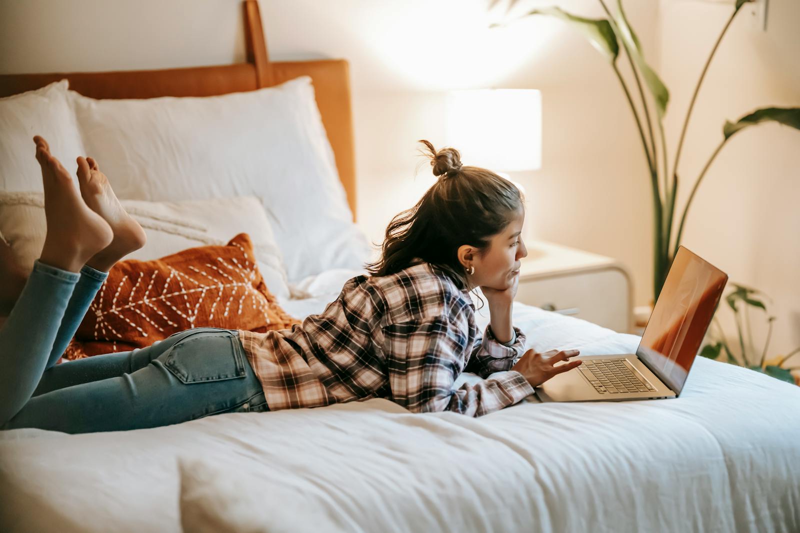 Side view of barefoot ethnic female with raised legs leaning on hand while browsing internet on netbook in bedroom